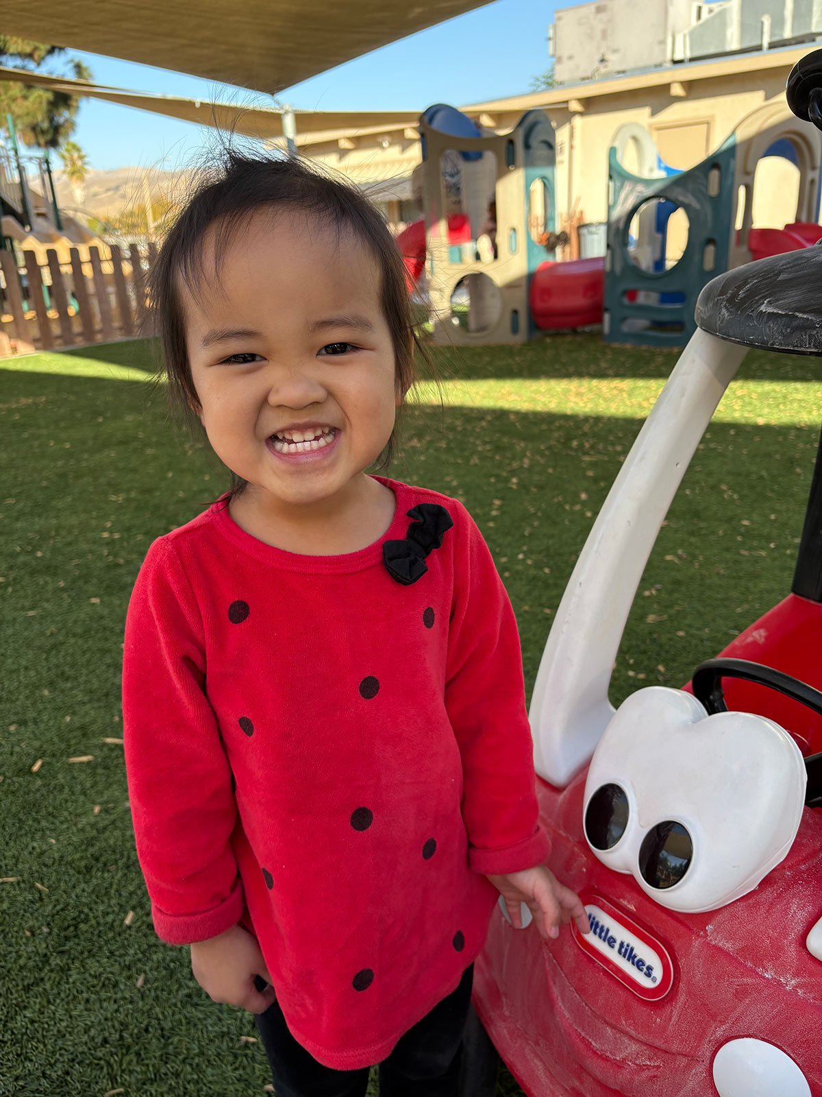 A smiling child in a red polka-dot shirt stands beside a red and white ride-on toy on artificial grass.